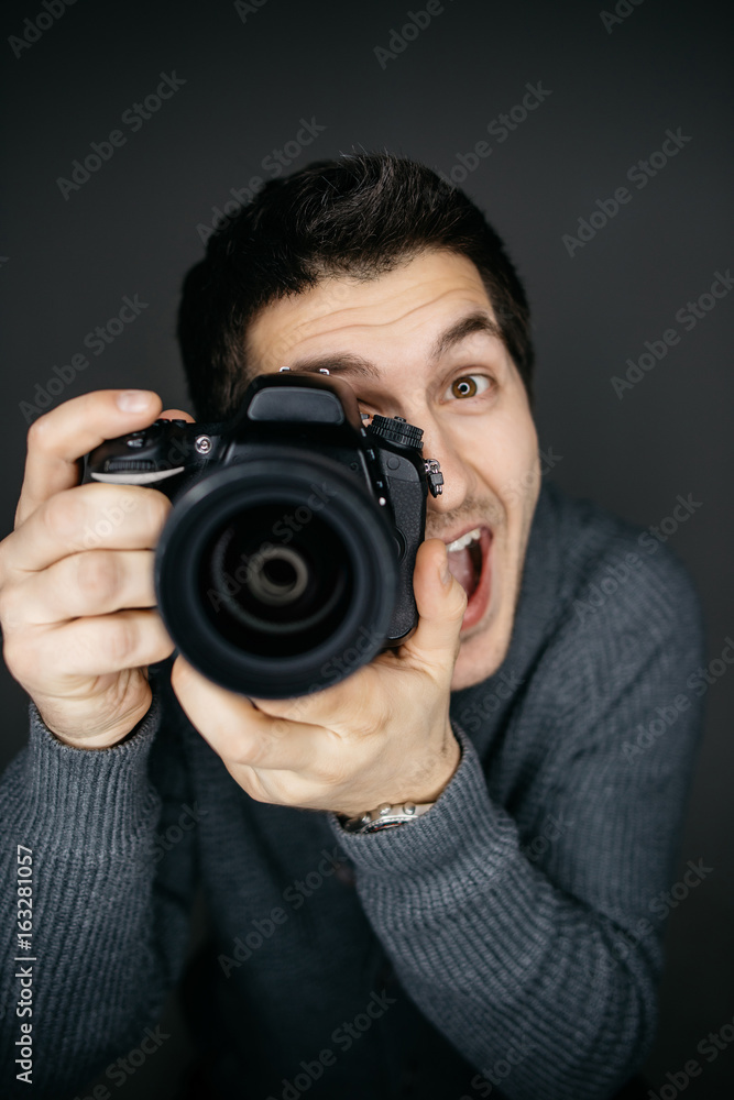 Young male photographer looking at the camera lens and smile. Funny expression. Studio portrait isolated on gray Young male photographer looking at the camera lens and smile. Funny expression. Studio portrait isolated on gray