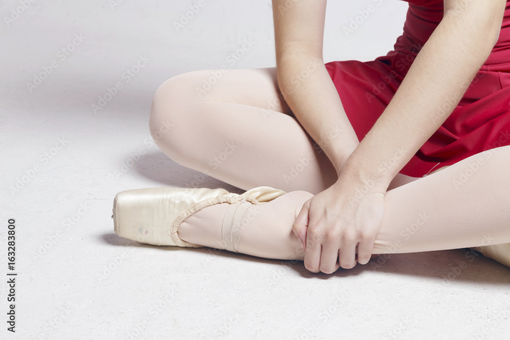 Fototapeta premium Ballerina sitting on a white floor, touching her foot, suffering pain on her fingers and ankle. No pain no gain.