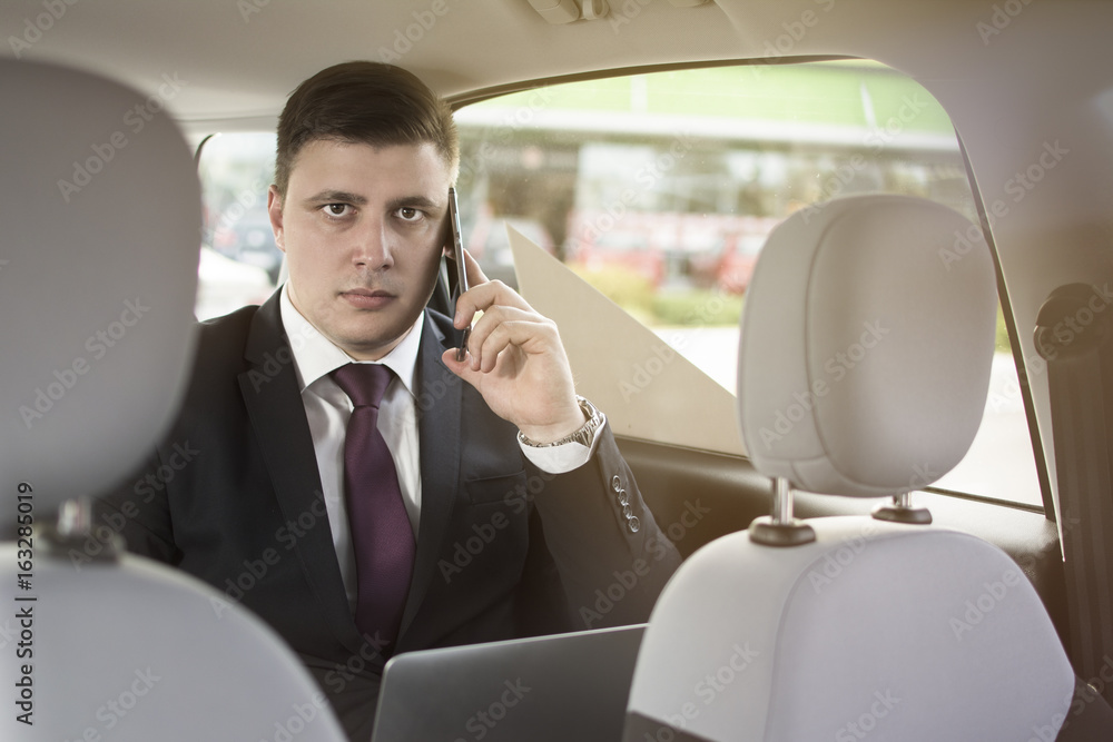 Businessman working while sitting in a car. A man is driven to work in his limo. Suit and tie businessman in the back seat making a call while the limo driver is driving. Man looking at the camera.