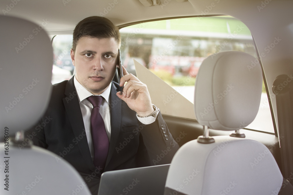Businessman working while sitting in a car. A man is driven to work in his limo. Suit and tie businessman in the back seat making a call while the limo driver is driving. Man looking at the camera. Businessman working while sitting in a car. A man is driven to work in his limo. Suit and tie businessman in the back seat making a call while the limo driver is driving. Man looking at the camera.