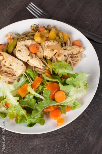  Whole wheat pasta with cooked chicken and vegetables including carrots, bell peppers, onion, and asparagus with a small salad as a side angled shot 