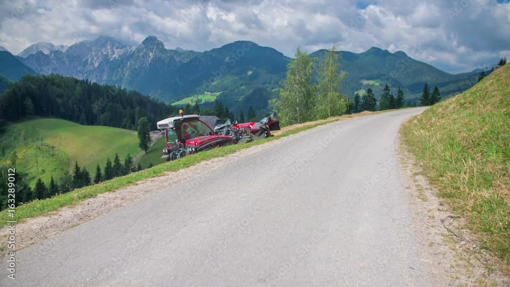 A small red tractor is driving back on a concrete road. A farmer finished preparing hay on a steep hill. The landscape is beautiful. A small red tractor is driving back on a concrete road. A farmer finished preparing hay on a steep hill. The landscape is beautiful.
