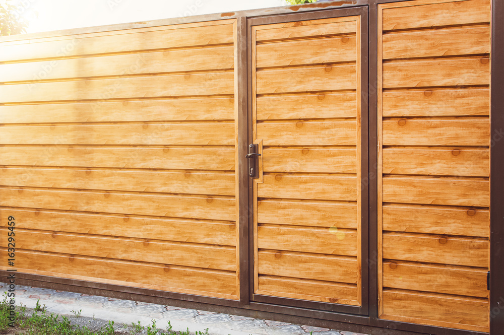 Fragment of a wooden brown modern fence with an entrance door In bright sunlight , closeup. Modern Style Design wooden Fence Ideas. Fragment of a wooden brown modern fence with an entrance door In bright sunlight , closeup. Modern Style Design wooden Fence Ideas.