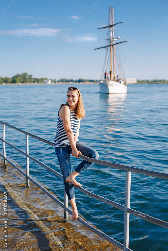 Portrait of white Caucasian blonde woman with tanned skin striped t-shirt and blue jeans sitting on pier by seashore lakeshore, with yacht boat ship on background on water, lifestyle summer