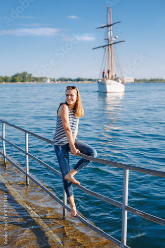 Portrait of white Caucasian blonde woman with tanned skin striped t-shirt and blue jeans sitting on pier by seashore lakeshore, with yacht boat ship on background on water, lifestyle summer