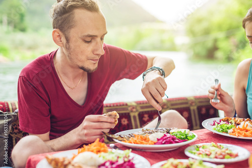 Young man eats vegetables and fish outdoors in a cozy restaurant on the water, discover new kitchen