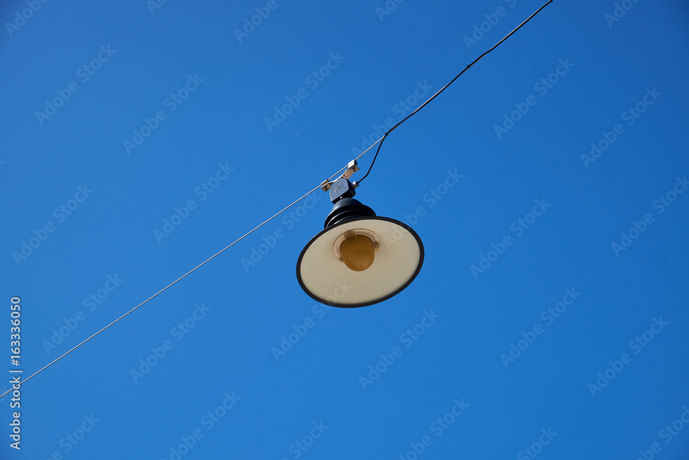 Round open street lamp hanging in a steel wire against a clear blue sky Round open street lamp hanging in a steel wire against a clear blue sky