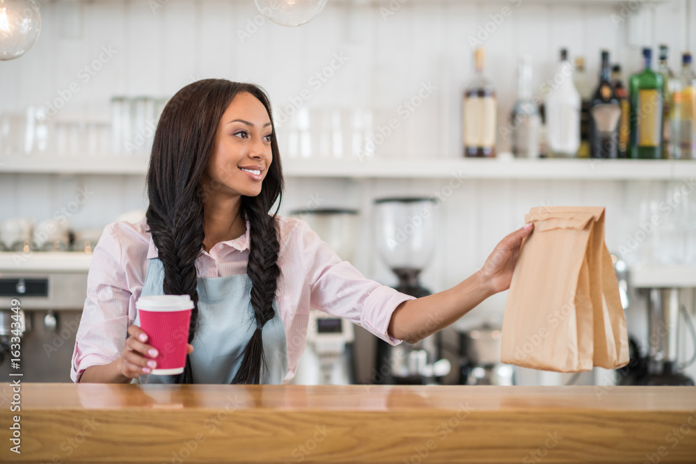 Obraz na plátně Smiling afro american waitress holding coffee to go and take away food in hands