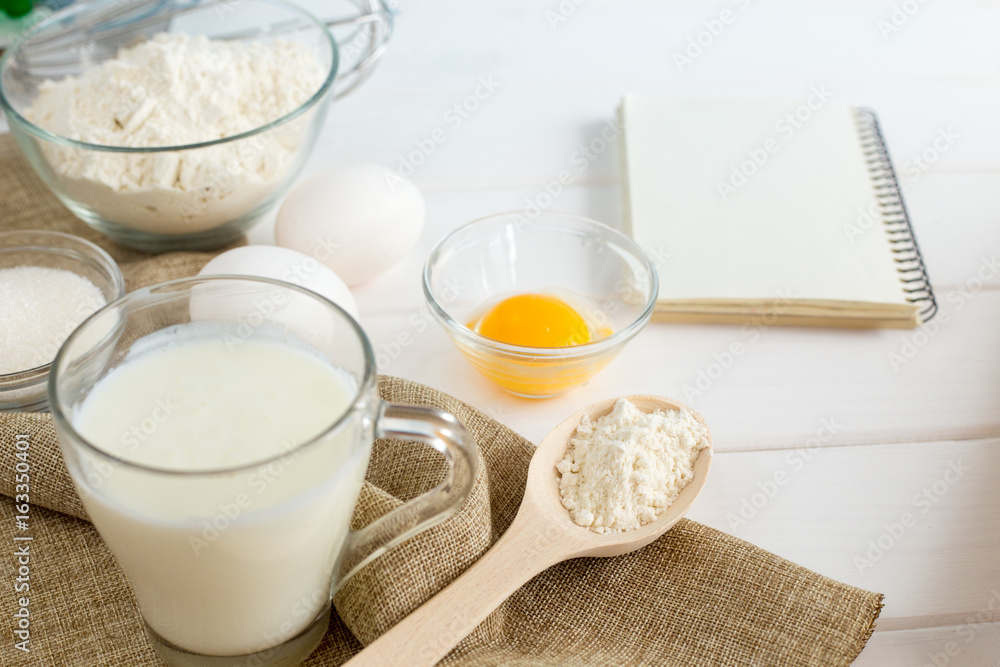 Baking in the kitchen - the ingredients of the test recipe (eggs, flour, milk, sugar) on a white wooden table with the inscription on top. Background layout with free text space. Baking in the kitchen - the ingredients of the test recipe (eggs, flour, milk, sugar) on a white wooden table with the inscription on top. Background layout with free text space.