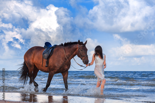 Girl in a white dress, leading a brown horse on the beach on a background of white clouds. The Baltic sea, Latvia.