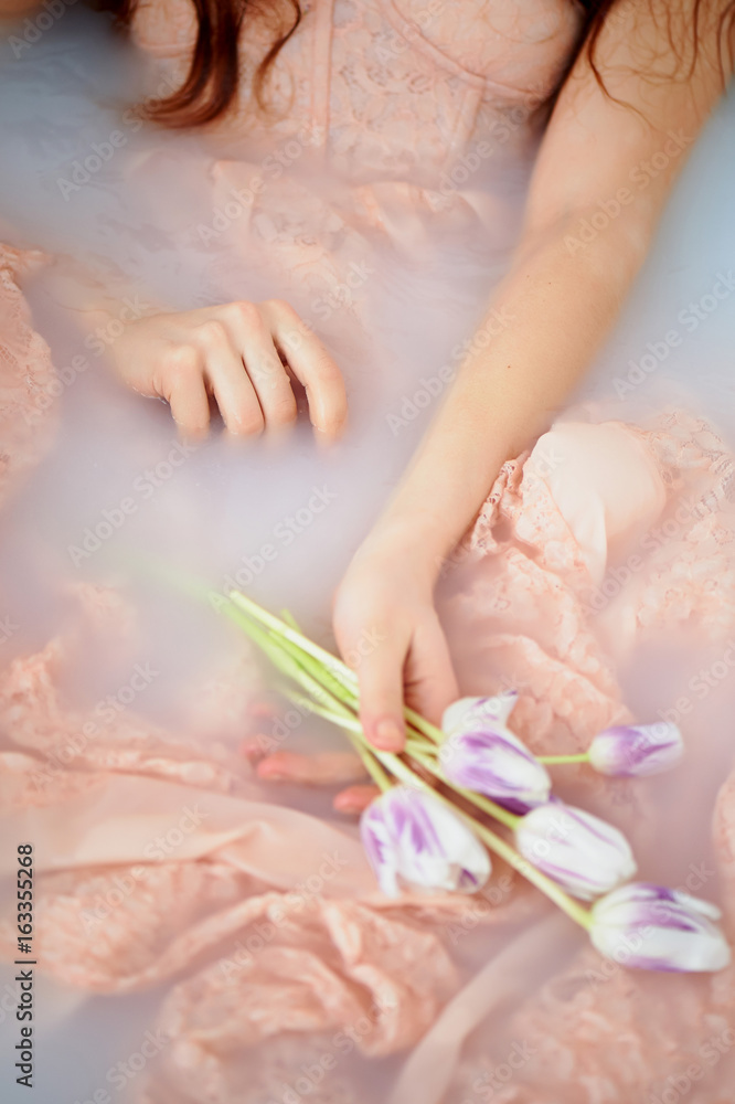 Beautiful romantic red-haired girl sitting in the bath and holding flowers .The face is not visible. In lace dress powdery pastel cream color