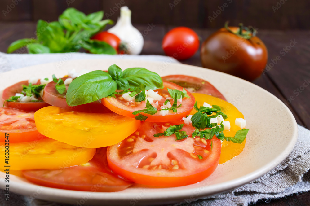 Spicy salad of yellow, red, black tomatoes, cut into circles with garlic and greens on a plate on a dark wooden background. Close up. Vegan cuisine.