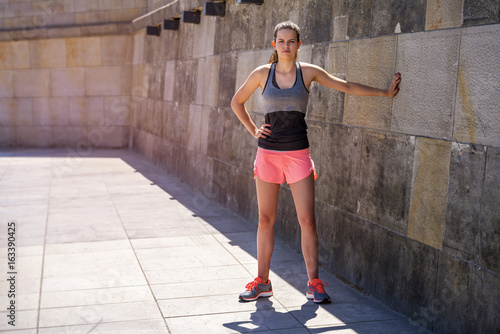 Young smiling female resting after an active fitness training while standing against wall, standing in full sun