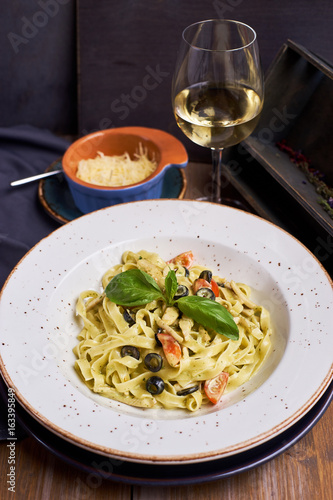 Pasta with chicken, olives and cheese on ceramic plate with glass of white wine on wooden table in rustic style.