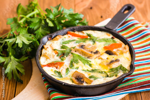 Frittata made from eggs, tomatoes, mushrooms, chicken and cheese in a frying pan and fresh parsley on a wooden table