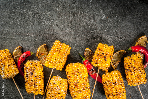 Summer food. Ideas for barbecue and grill parties. Grilled corn grilled on fire. With a sprinkle of cheese (mexican elotes), hot chili pepper and lemon. On a dark stone table. Top view copy space
