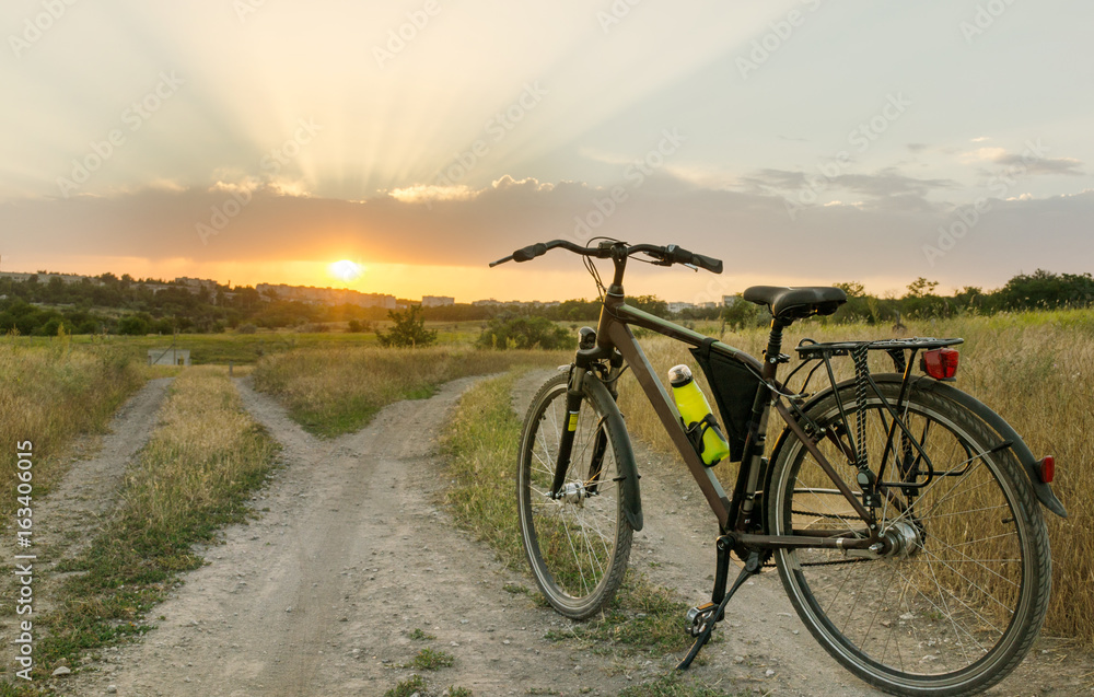Obraz premium The bicycle stands on a village road that is divided into two parts. Sunset time.