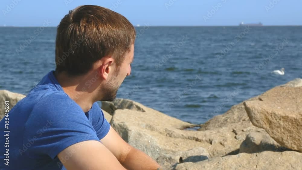 Happy young male sitting on stones at embankment and enjoying beautiful seascape Happy young male sitting on stones at embankment and enjoying beautiful seascape