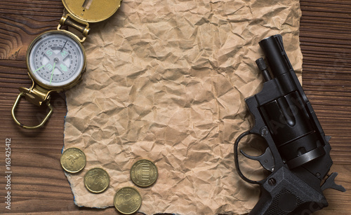 Adventurer, western, reward, treasure hunt concept. Compass, revolver gun, money and blank crumpled brown page paper on wooden table.