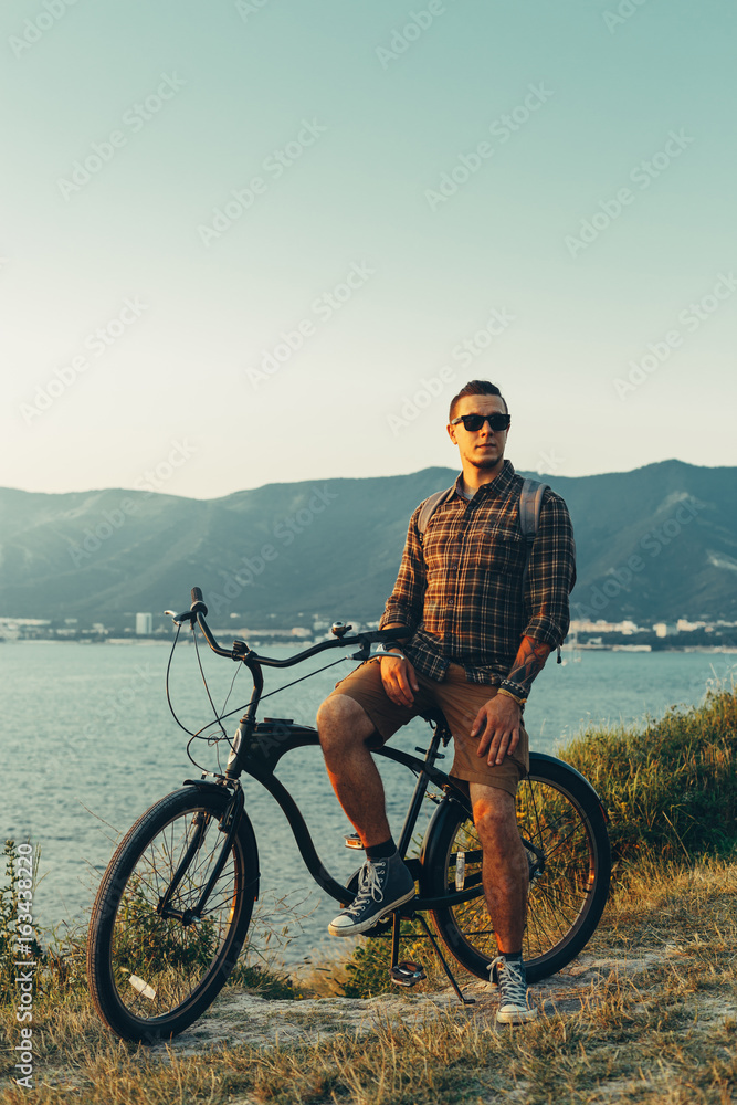Fototapeta premium Handsome Young Man Standing With Bike On Coast And Enjoying View of Nature Sunset Vacation Traveling Relaxation Resting Concept