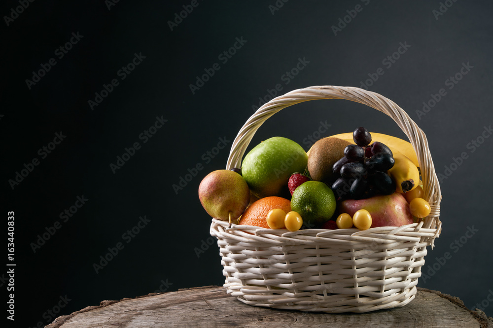 Assortment of fresh raw fruits in wicker basket on wooden stump isolated on black background. Healthy eating concept.