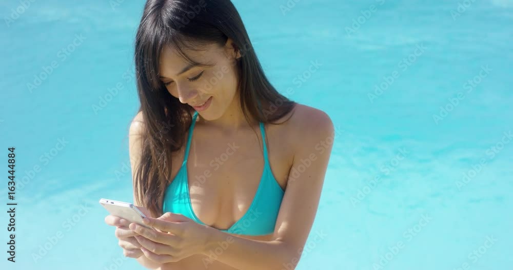 Attractive young woman sending a text message on her mobile phone as she relaxes poolside in the hot summer sun Attractive young woman sending a text message on her mobile phone as she relaxes poolside in the hot summer sun