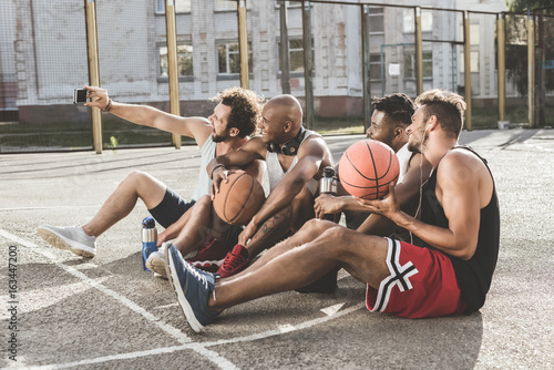 multiethnic group of men taking selfie on smartphone while sitting on court after basketball game