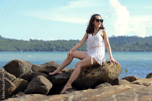 beautiful woman wear sunglasses posing sit on the rock in the sea beach with blue sky