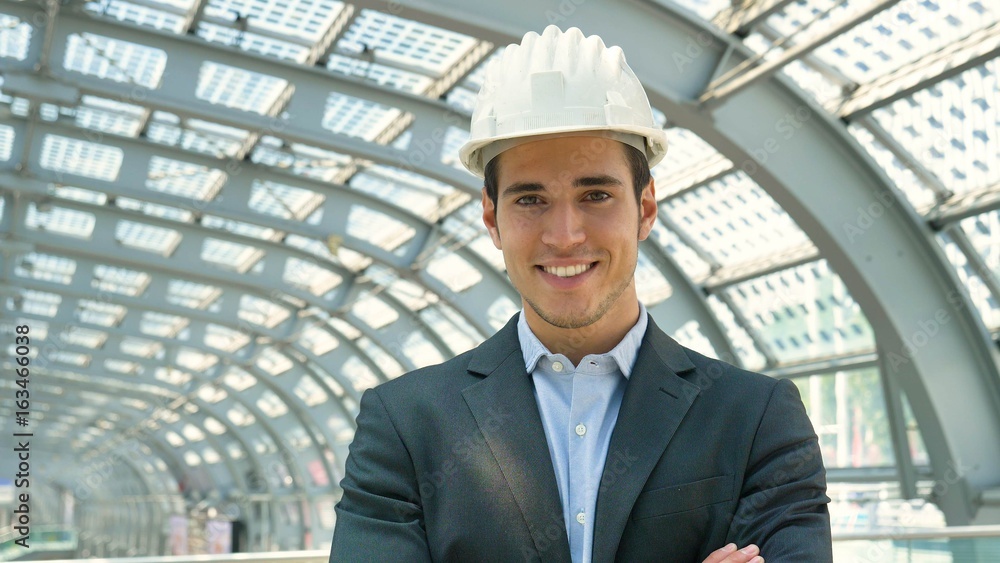 A young handsome, engineer, foreman, architect (builder) looks at the construction of the structure, at the station, wearing a white protective helmet, smiling in a suit. Concept: good work, project. A young handsome, engineer, foreman, architect (builder) looks at the construction of the structure, at the station, wearing a white protective helmet, smiling in a suit. Concept: good work, project.