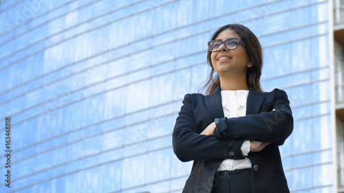 Portrait of young beautiful business woman (student) in suit, glasses, smiling, successful looking at sides, with skyscraper background. Concept: new business, communication, Arab, banker, manager.
