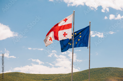 The European Union and Georgian flags fly beside each other, on a background mountain and sky .