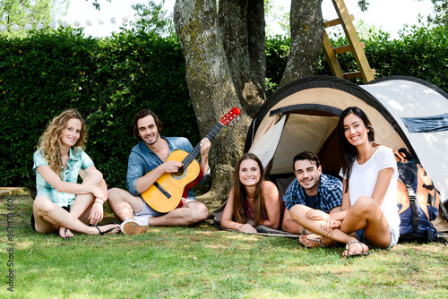 group of five young people playing guitar in a camp site during summer holiday vacation adventure trip