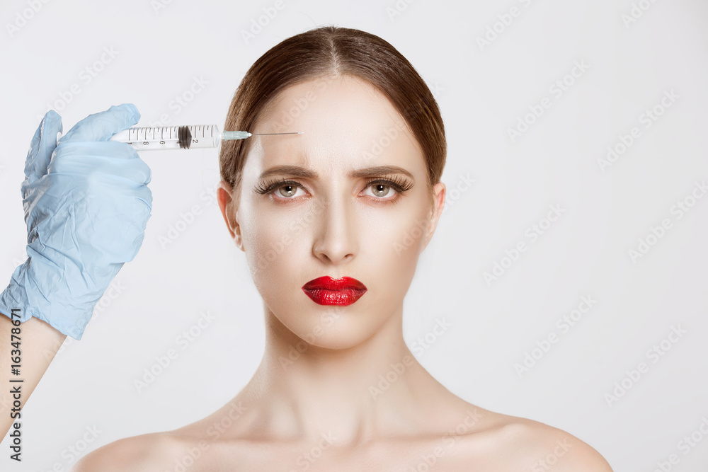 Portrait of beautiful worried young woman afraid of anti aging serum shot in forehead wrinkles plastic surgery doctor's hand blue gloves holding injection over woman's face. Negative face expression Portrait of beautiful worried young woman afraid of anti aging serum shot in forehead wrinkles plastic surgery doctor's hand blue gloves holding injection over woman's face. Negative face expression