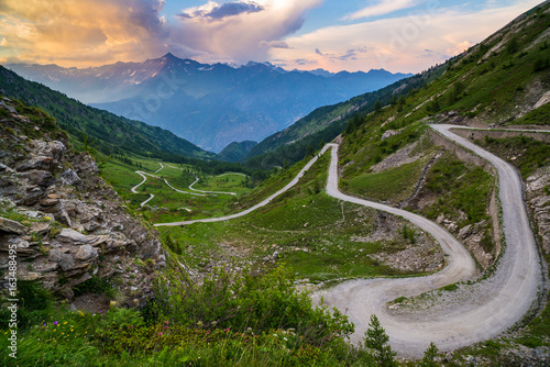 Dirt mountain road leading to high mountain pass in Italy (Colle delle Finestre). Expasive view at sunset, colorful dramatic sky, adventures in summer time, Italian Alps.
