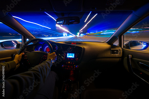 Hands on the wheel the car moves with fast speed on the highway at night. Blurred road with lights with a car at high speed. © narozhnii