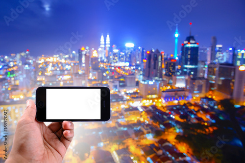 Hand holding a smartphone with Blur image of Kuala Lumpur city at night at background.