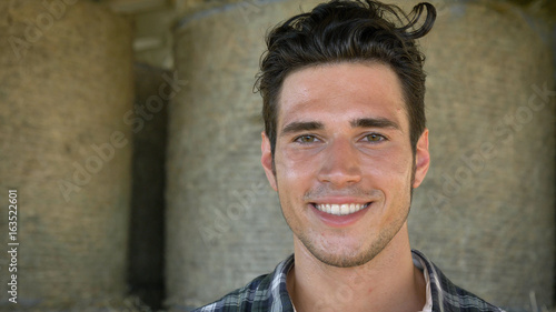 Portrait of a handsome young farmer (student) sitting on haystack, beautiful smile, in shirt, happy smiling. Concept: clean air, ecology, agriculture, hay, farmers, successful people, business.