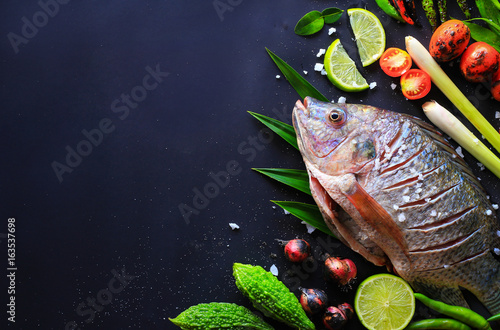 Top view delicious fresh fish and herbs and spices, lemon, tomato, chili and various vegetables on dark wood background, space for texture. Healthy food, diet or cooking concept.