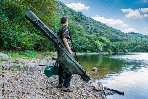 Fishing adventures, carp fishing. Fisherman on a lake shore with camouflage fishing gear, green bag and mimetic rod holdall