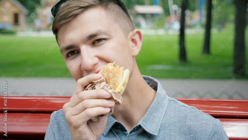 A man with a delight eating a hamburger sitting on a bench in a city park A man with a delight eating a hamburger sitting on a bench in a city park