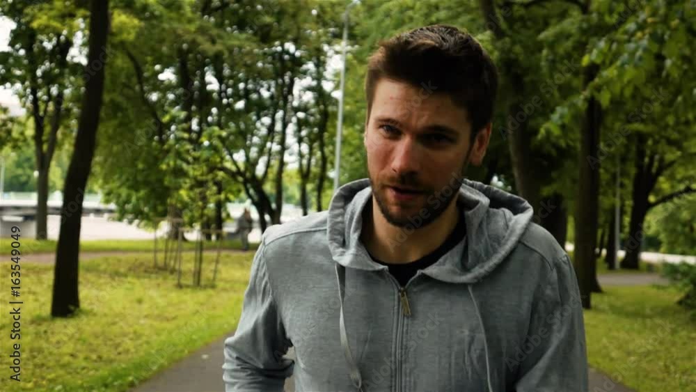 Smiling young attractive powerful athlete man run in the park in gray jacket. He tired. Green trees background. Extreme slow motion footage. Smiling young attractive powerful athlete man run in the park in gray jacket. He tired. Green trees background. Extreme slow motion footage.
