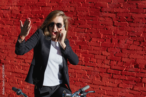 Young beautiful fashionable Caucasian lady on bicycle wearing formal suit having expressive phone conversation in front at red brick wall background. People, technology and active lifestyle concept.
