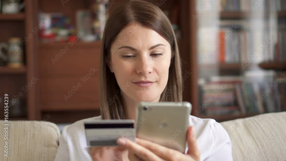 Slow Motion Woman in white blouse sitting on sofa in living room buying online with credit card on smartphone Slow Motion Woman in white blouse sitting on sofa in living room buying online with credit card on smartphone