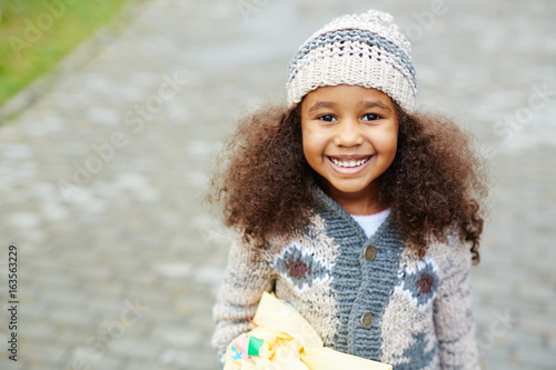 Portrait of cute African-American girl looking up at camera and smiling wearing knit hat and sweater on warm autumn day photo