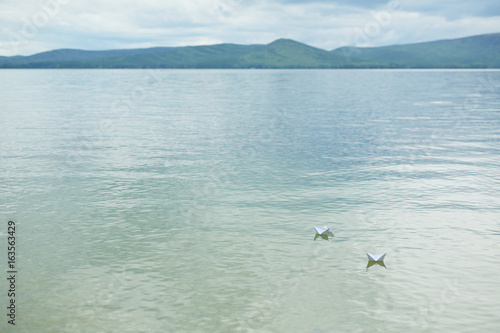 Two paper ships floating on calm water of clear blue lake with mountain silhouette in background