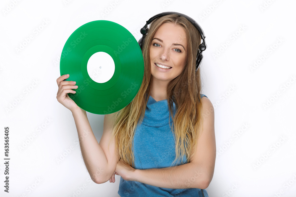 Happy young woman in big black professional dj headphones holding trendy green colorful vinyl record posing against white studio backdrop Happy young woman in big black professional dj headphones holding trendy green colorful vinyl record posing against white studio backdrop