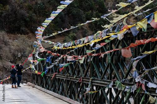 Couple tourists with tear colorful Tibetan prayer flags waving and swaddled with bridge over frozen river at Thangu and Chopta valley in winter in Lachen. North Sikkim, India. photo