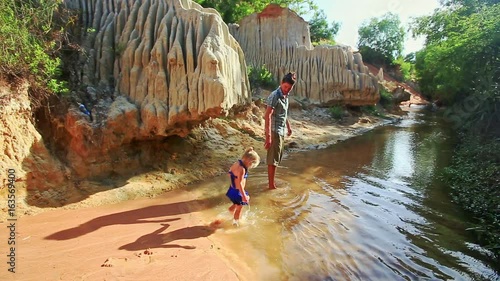 Little girl with pigtail in blue swimsuit plays on wet sand beach walks with dad in stream water on sunny morning