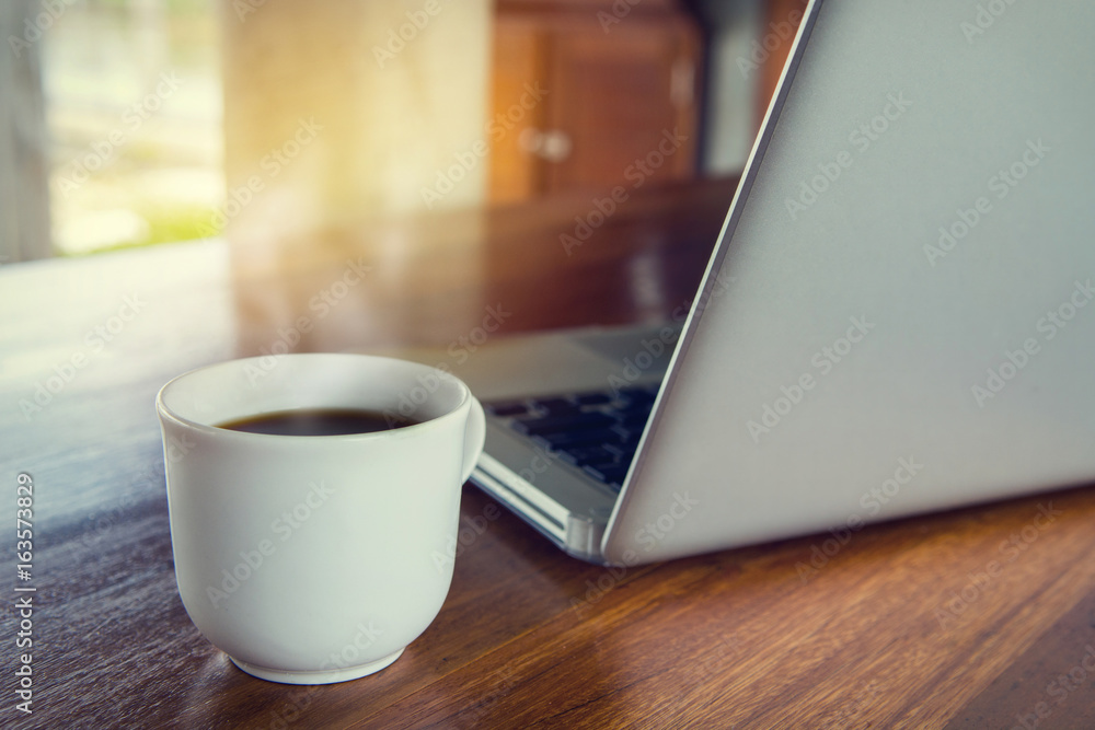 Selective focus of Cup of coffee and a laptop on dark wooden with sunshine background.