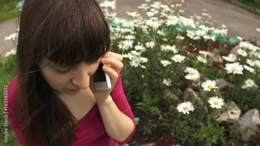 Young brunette woman in pink blouse calling on white phone while sitting at blooming daisies in city park on a summer day.